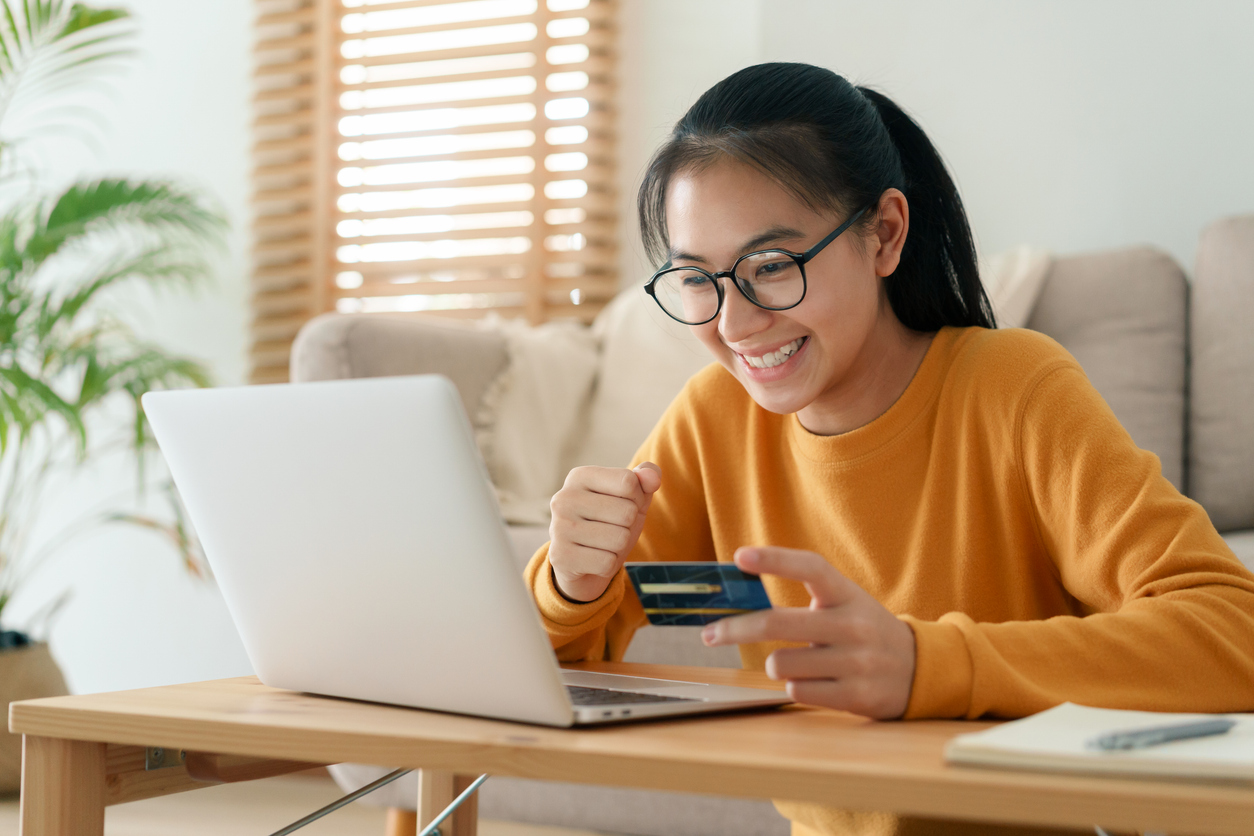 young woman smiling in front of computer in her living room holding a credit card